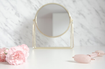 Jade roller and pink flowers with golden mirror on white marble background.