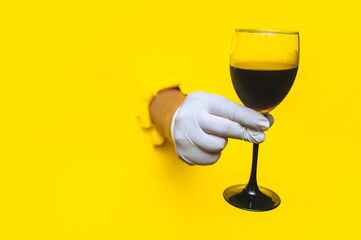 From a torn hole in bright yellow paper, a right doctor's hand appears in a white latex glove with a glass of dark red wine. The concept of the benefits of alcohol, drunkenness and hangover.Copy space
