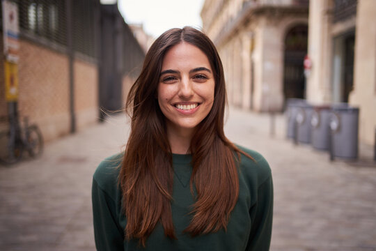 Close up portrait joyful beautiful young Caucasian gen z woman happy smiling on city street. Female people with nice expression looking at camera in open air. Spanish girl posing for picture outdoor