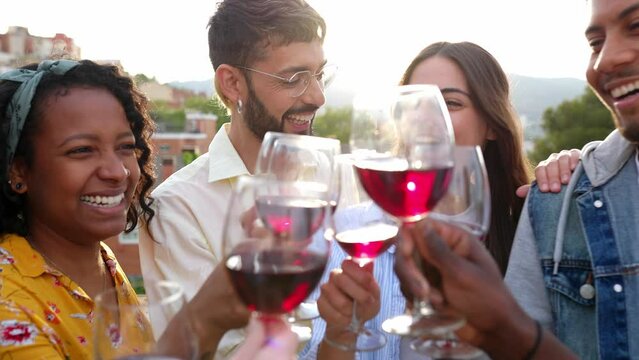 Group of multiracial young friends hanging out while having party at rooftop terrace. Millennial people enjoying time together toasting red wine on summer celebration. Friendship and youth concept.