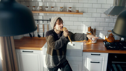 High angle shot of carefree young man with beanie singing into a ladle like a microphone and freely dancing to music in a kitchen at home	
