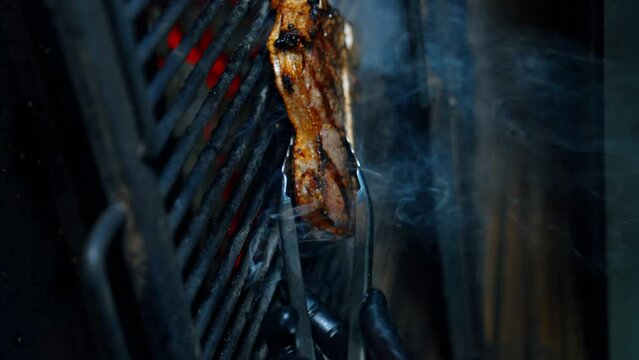 Meat on the bone is cooked on the grill. Gloved hand turns the piece of beef with tongs. Vertical screen. Close up.