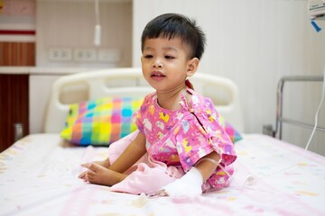 2-year-old baby dressed in pink patient clothes sitting on bed at the hospital.