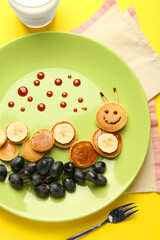Creative serving for kids. Plate with cute caterpillar made of pancakes, grapes and banana on yellow background, flat lay