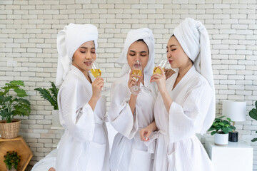Three young women wearing bathrobes holding wine glasses stand talking in a bedroom