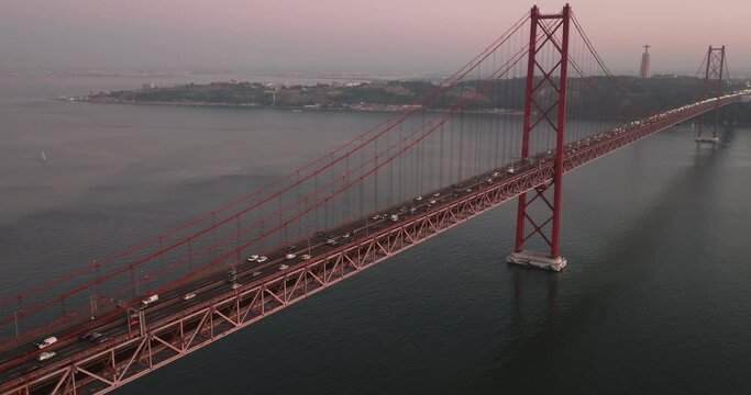Aerial drone shot towards the 25th April Bridge, Ponte 25 de Abril, and Cristo Rei in Lisbon, Portugal, Europe. Dusk after sunset with pink and orange sky. Drone orbiting. Shot in ProRes 422 HQ