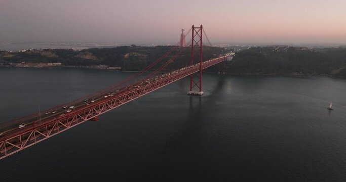 Aerial drone shot towards the 25th April Bridge, Ponte 25 de Abril, and Cristo Rei in Lisbon, Portugal, Europe. Dusk after sunset with pink and orange sky. Flying forwards. Shot in ProRes 422 HQ