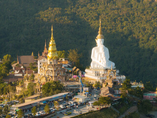 The setting sun illuminates a striking white Buddha statue beside an ornate temple, with a lively street scene unfolding below.