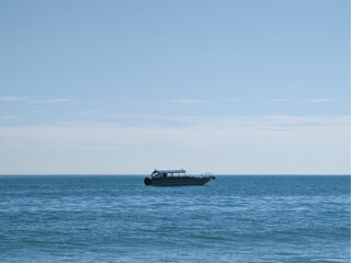 A serene and calm seascape under a clear sky. A single boat, white in color with a canopy, is floating peacefully on the water. The sea has gentle waves, indicating calm weather conditions.