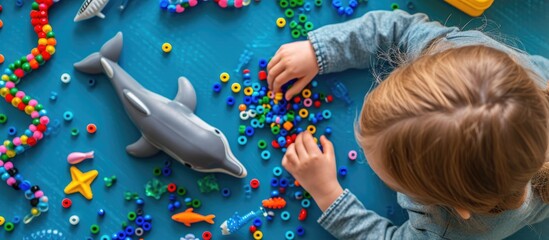 Child developing creativity and fine motor skills by crafting a dolphin from beads. Overhead perspective. - obrazy, fototapety, plakaty