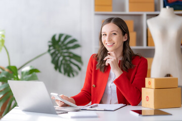 Smiling entrepreneur in a red blazer organizing product shipments in her startup's bright and...