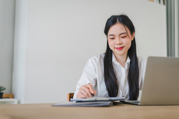 Focused asian professional businesswoman calculating budget using laptop and taking notes in a bright office environment.