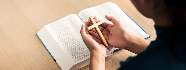 Female god believer holding wooden cross on opened holy bible book at light wooden church table....