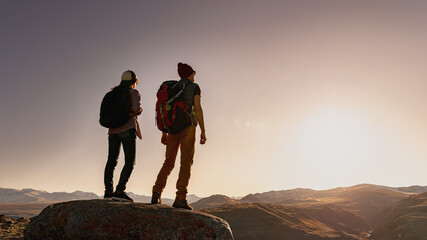 Two young active silhouettes of hikers with backpacks are standing on big rock and looks at sunset...