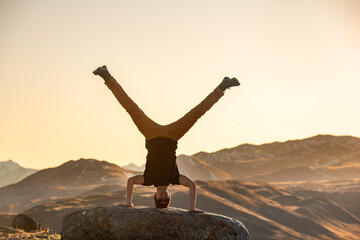 Active young man is standing upside down on big rock in sunset mountains - obrazy, fototapety, plakaty