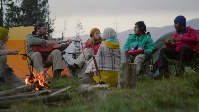 Diverse hiking buddies eat and sit near campfire in camp. Caucasian man plays guitar and talks with African American teenager. Group of tourists resting in campsite in forest. Outdoor activities.