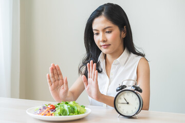 Intermittent fasting with clock, asian young woman dieting hand refuse waiting time to eat low carb food, green vegetable salad on plate. Eat food healthy first meal on brunch, lunch on table at home.