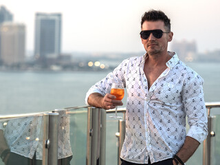 Stylish man with a glass of drink standing on the empty deck of a cruise liner against the backdrop...