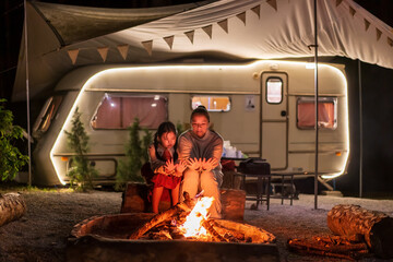 Asian family mother and child camper or kid girl travel camping sitting together on log to warming themselves by bonfire at night with campervan or RV motorhome in holiday at Doi Bo Luang Forest Park - obrazy, fototapety, plakaty