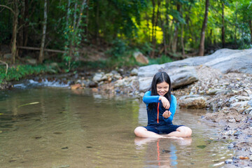 Asian child or kid girl sitting smiling playing in nature water stream and garden forest at phalad waterfall in lan sang national park at Tak Thailand for summer holiday relax and travel to happy fun