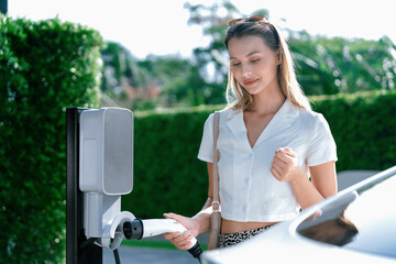 Young woman travel with EV electric car charging in green sustainable city outdoor garden in...