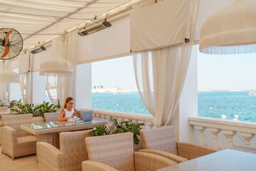 Woman coffee cafe laptop sea. Female freelancer using laptop on coffee break in modern cafe with sea view in the morning