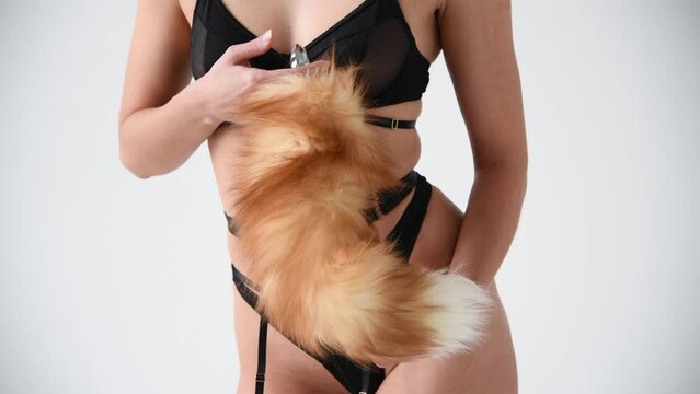 Close-up view of slim caucasian woman wearing black lingerie and holding butt plug with orange fox tail next to belly against white background. Soft focus. Real time video. Adult sex toys theme.