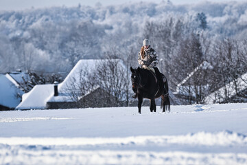 A young red-haired woman rides her horse at a walk across a Schebeckte meadow with a snowy village...