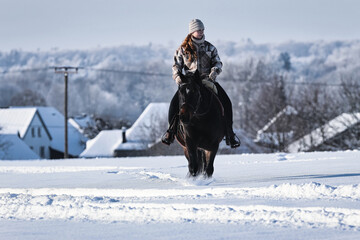 A young red-haired woman rides her horse at a walk across a Schebeckte meadow with a snowy village...