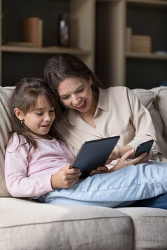 Vertical shot pretty woman and daughter sit on sofa at home with digital tablet gadget, spend free time on internet, buying via electronic commerce marketplace app, enjoy funny videos and application