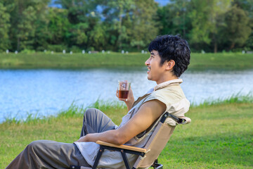 Asian male man sitting on chair, drinking coffee and feeling relax during camping beside lake and mountain.