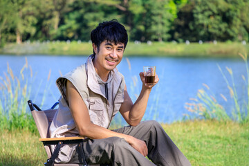 Young Asian male man sitting on chair, drinking coffee, feeling relax and raising hands, during camping beside lake and mountain. Looking at camera.