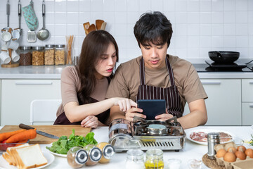 Portrait of young Asian couple male man female woman cheerful sitting and preparing food for meal, enjoy cooking, in kitchen at home. Reading recipe in tablet.