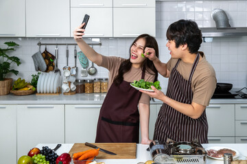 Portrait of young Asian couple male man female woman cheerful standing and preparing food for meal, enjoy cooking, in kitchen at home. Taking selfie.
