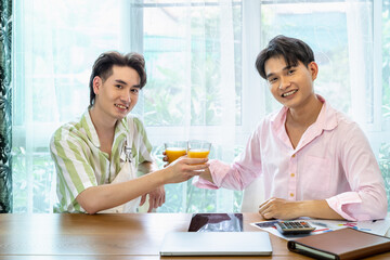 Cute Asian LGBT gay man couple, spend time together, with tablet, and laptop, on table. Holding orange juice glass.  Healthy lifestyle and work from home. Looking at camera.