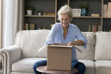 Happy attractive elderly woman spend time at home sitting on sofa, open parcel box, smiling, get...