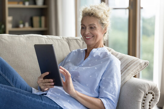 Smiling attractive mature female in casual clothes resting on sofa with digital tablet enjoy easy and comfort usage of gadget for fun, leisure or hobby, buying goods and services on internet at home