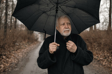 An elderly man in a black coat with an umbrella stands outside in the rain, looks to the side and holds his collar