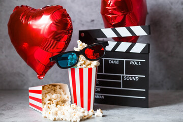 A banner for the film industry. A romantic movie date. A movie camera, 3D glasses, popcorn and foil balloons in the shape of hearts on a gray background. The film will premiere on Valentine's Day.