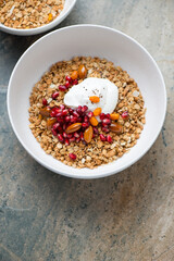 White bowl with granola, honey, pomegranate, nuts and yogurt, vertical shot on a beige granite background, elevated view