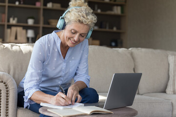 Smiling mature woman in wireless headphones make notes, studying online, writing information in copybook, looking interested, enjoy e-learn, gain new knowledge on retirement use modern tech. Education