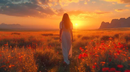 Poster  a woman standing in a field of flowers with the sun setting behind her and a mountain range in the distance, behind her is a field of red poppies. © Olga