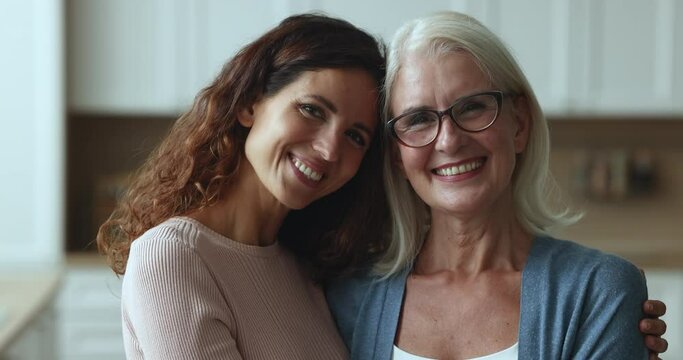 Happy senior mom and adult daughter woman posing for family portrait at home, looking at camera with toothy smiles, laughing, hugging, standing close with faces touch, enjoying warm relationship