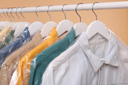 Dry-cleaning service. Many different clothes in plastic bags hanging on rack against beige background, closeup