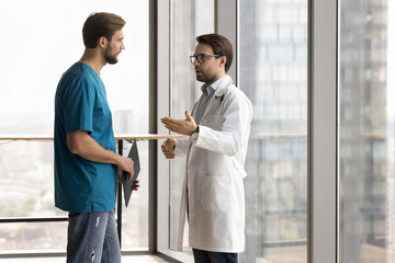 Serious doctor talking to hospital nurse man, giving instructions. Medical colleagues in white and...