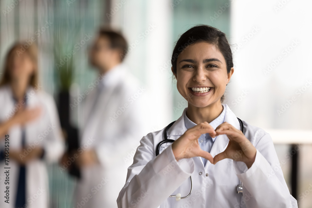 Wall mural cheerful young indian doctor woman making hand heart, looking at camera with happy toothy smile, sho