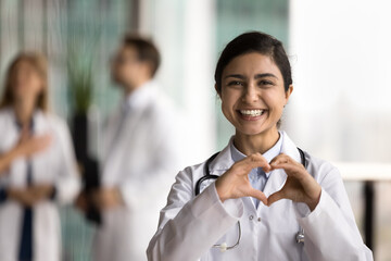 Cheerful young Indian doctor woman making hand heart, looking at camera with happy toothy smile, showing romantic gesture of love, care, health protection, cardiology healthcare