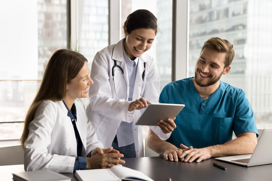 Cheerful diverse clinic medical colleagues in uniforms using tablet computer at workplace together, talking, laughing, discussing healthcare online service, enjoying modern Internet technology