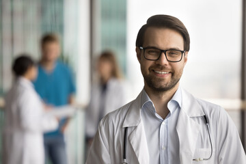Happy confident doctor man in glasses and white coat looking at camera in linic space, posing for front portrait with colleagues talking in blurred background, smiling for head shot
