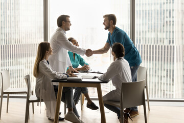 Two male doctors thanking each other for collaboration, shaking hands over table at multiethnic team meeting, talking, smiling, laughing in indoor office space with large window and city view - obrazy, fototapety, plakaty
